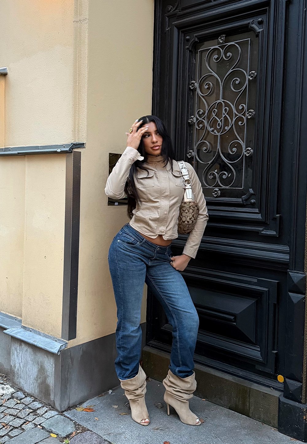 Bėriniuotos smėlio spalvos trumpa striukė, melsvi džinsai ir smėlio spalvos laisvo stiliaus aulinukai su kulnais. Stovi prie juodų kalviškų durų su dekoratyvinėmis detalėmis.