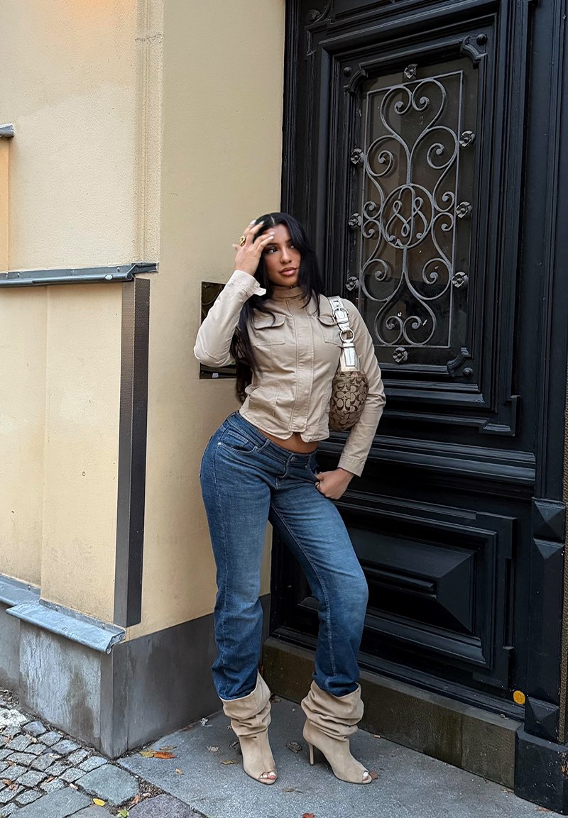 Giacca beige corta, jeans di denim blu e stivali beige con tacco morbido. In piedi accanto a una porta nera in ferro battuto con dettagli decorativi.