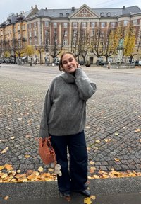 Pulover gri, supradimensionat, cu guler înalt, asortat cu blugi negri, cu croială largă. Ținând o geantă texturată, de culoare ruginie, cu un pandantiv.