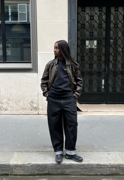 Chaqueta bomber de cuero marrón, sudadera negra con logo amarillo, vaqueros negros holgados con puños, zapatillas negras con acentos rojos, de pie en la acera.