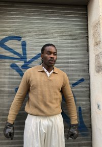 Un homme portant un pull beige, un pantalon blanc plissé et des gants noirs se tient devant un volet métallique avec un graffiti bleu.