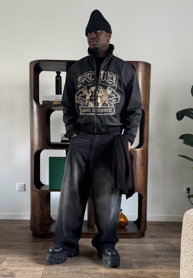 Homme portant un bonnet noir, une veste en cuir graphique, un pantalon large noir et des chaussures compensées, debout à l'intérieur près d'une étagère en bois en tenant une écharpe noire.