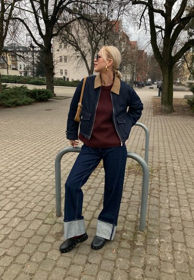 Chaqueta de mezclilla con un cuello beige, suéter burdeos, jeans oscuros de pierna ancha con puños grises y zuecos negros, en un entorno al aire libre.