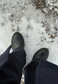 Άτομο που φοράει μαύρα μποτάκια και σκούρο παντελόνι, στέκεται σε έδαφος που είναι μερικώς καλυμμένο με χιόνι, βρωμιά και διάσπαρτα κλαδ άκια.