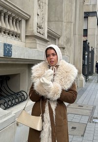 Femme portant un manteau en peau retournée marron, un bonnet en tricot et des moufles, tenant un sac à main beige, debout sur un trottoir en ville près d'un bâtiment en pierre.