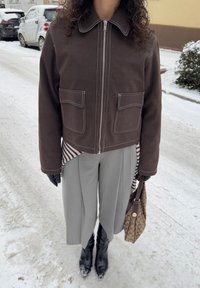 Veste marron à fermeture éclair avec surpiqûres blanches et deux poches avant, associée à un pantalon plissé gris et des bottes noires montantes. Sac marron à motifs.
