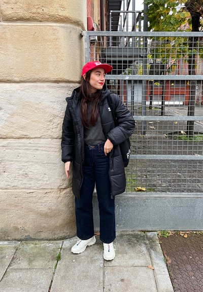 Abrigo negro acolchado con capucha sobre un suéter gris, combinado con jeans oscuros. Una gorra roja y zapatillas blancas completan el conjunto. El fondo presenta una valla.