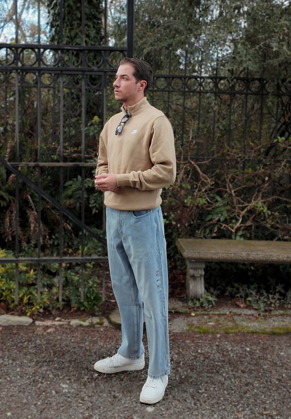 Joven con sudadera beige y vaqueros azul claro de pie junto a una valla de metal negra y un banco de piedra en un jardín.