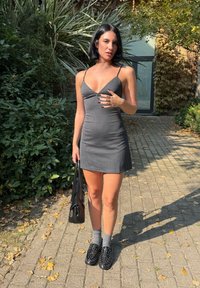 Gestreiftes Mini-Kleid in Schwarz-Weiß mit Spaghetti-Trägern und V-Ausschnitt, kombiniert mit grauen Socken und genoppten schwarzen Loafers, während sie eine schwarze Tasche hält.