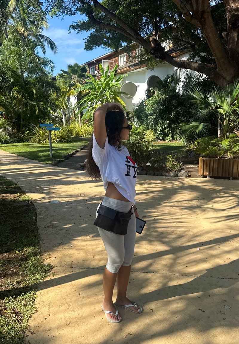 Femme portant des lunettes de soleil et des vêtements décontractés se tenant sur un chemin ensoleillé, entourée de plantes tropicales et d’arbres près d’un bâtiment blanc.