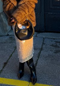 Sac hobo rembourré noir de forme arrondie, porté avec un manteau marron en fausse fourrure, superposé à une jupe blanche transparente ornée de sequins et des bottes noires à bouts pointus.