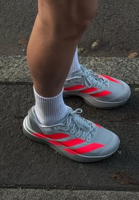 Fehér zoknit és szürke futócipőt viselő lábak, élénk piros csíkkal, aszfalt úttesten állnak egy járdaszegély közelében.