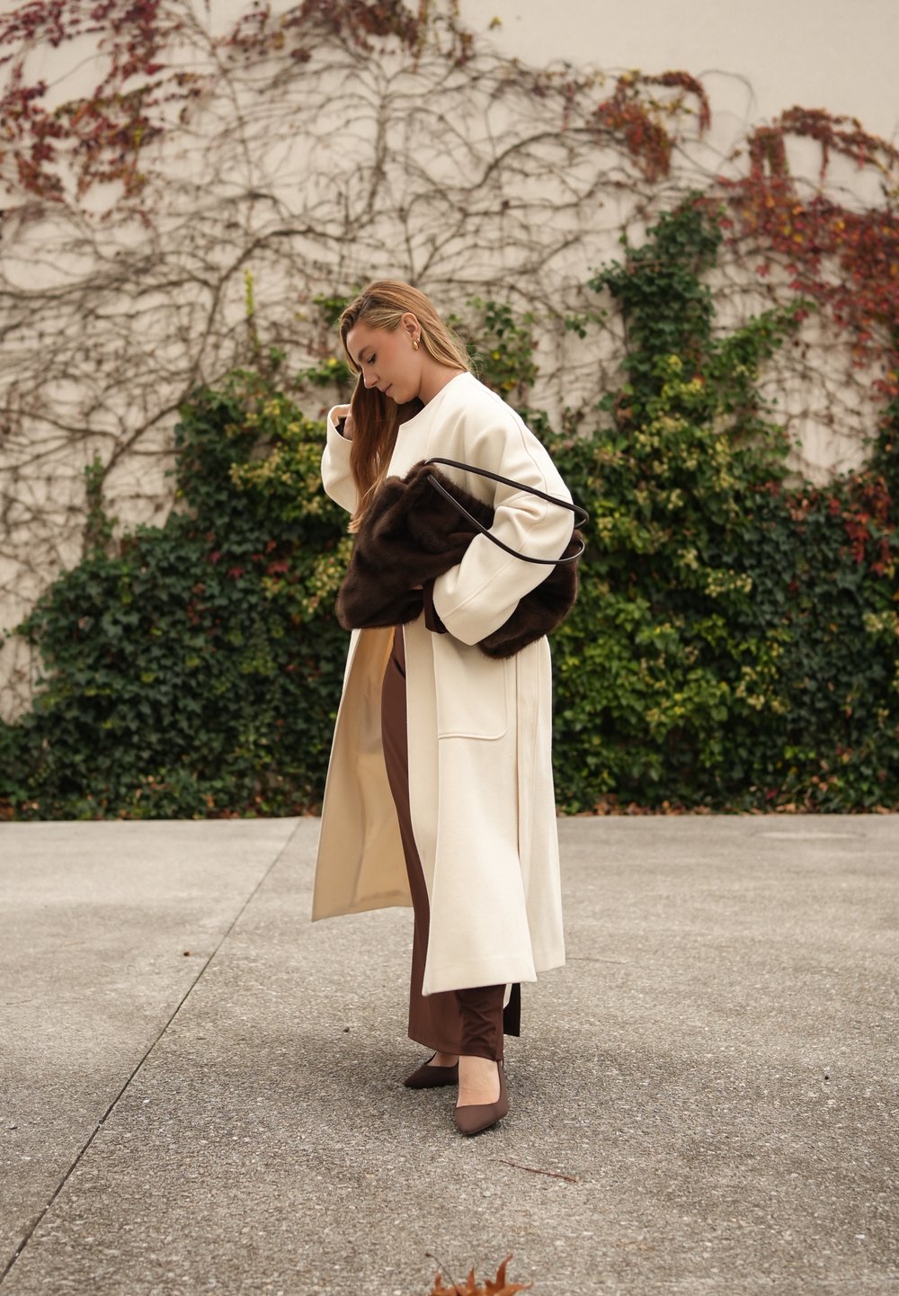 Długi kremowy płaszcz, brązowe dopasowane spodnie i brązowe szpilki. Trzyma ciemnobrązową futrzaną torebkę. Gładkie tekstury, minimalistyczny design i neutralne kolory.
