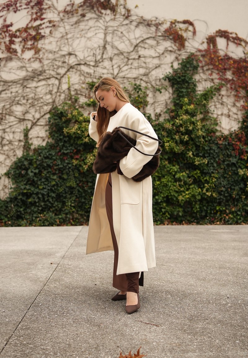 Langer cremefarbener Mantel, braune, eng anliegende Hosen und braune Absätze. Hält eine dunkelbraune Pelztasche. Glatte Texturen, minimalistisches Design und neutrale Farben.