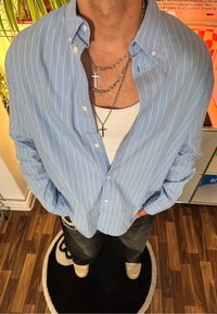 Camicia button-up a righe blu chiaro con una vestibilità comoda, indossata sopra una canotta bianca, decorata con molte collane in argento.