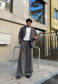 Homme aux cheveux courts et bouclés portant un costume gris surdimensionné, une chemise blanche et des lunettes, debout sur un trottoir carrelé en milieu urbain.