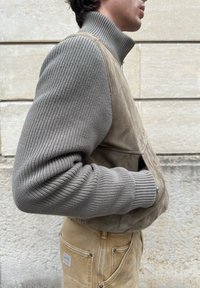 Άνδρας που φοράει γκρι ριπ πλεκτό πουλόβερ με ψηλό γιακά και μπεζ σουέτ γιλέκο, στέκεται μπροστά σε πέτρινο τοίχο.