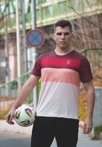 Sportinė marškinėliai su horizontaliomis raudonomis, rožinėmis ir baltomis juostelėmis. Lengvas audinys. Modelis laiko futbolo kamuolį, dėvi juodus kelnes.