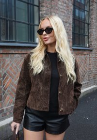 Casaco de camurça castanha com botões dourados, combinado com uma camisola preta e calções de couro. Cabelo loiro, comprido e ondulado. Fundo urbano de tijolos.