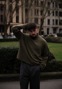 Pull à col roulé en maille côtelée vert olive avec une coupe ample, associé à un pantalon gris cintré. Cadre extérieur avec de la verdure et des bâtiments.