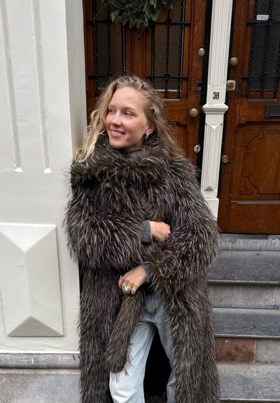 Abrigo largo de piel sintética tupida en tonos gris oscuro y marrón, con un cuello ancho, llevado sobre un suéter gris y jeans de mezclilla clara.