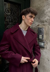 Manteau en laine bordeaux avec larges revers, noué à la taille, porté sur un pull en maille gris clair, devant un mur en pierre texturé et un interphone.