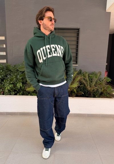Hombre con sudadera verde "QUEENS", pantalones cargo azul oscuro, zapatillas blancas y gafas de sol, de pie junto a plantas y pared gris.
