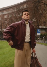 Veste en cuir bordeaux portée sur un pull en maille torsadée crème, associée à un pantalon beige et tenant un sac en cuir marron. Fond urbain.