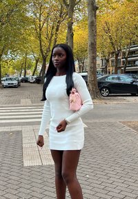 Nainen valkoisessa mekossa kävelee kaupunkikävelytiellä lähellä suojatietä, kantaen vaaleanpunaista käsilaukkua, taustalla syksyisiä puita ja parkkeerattuja autoja.