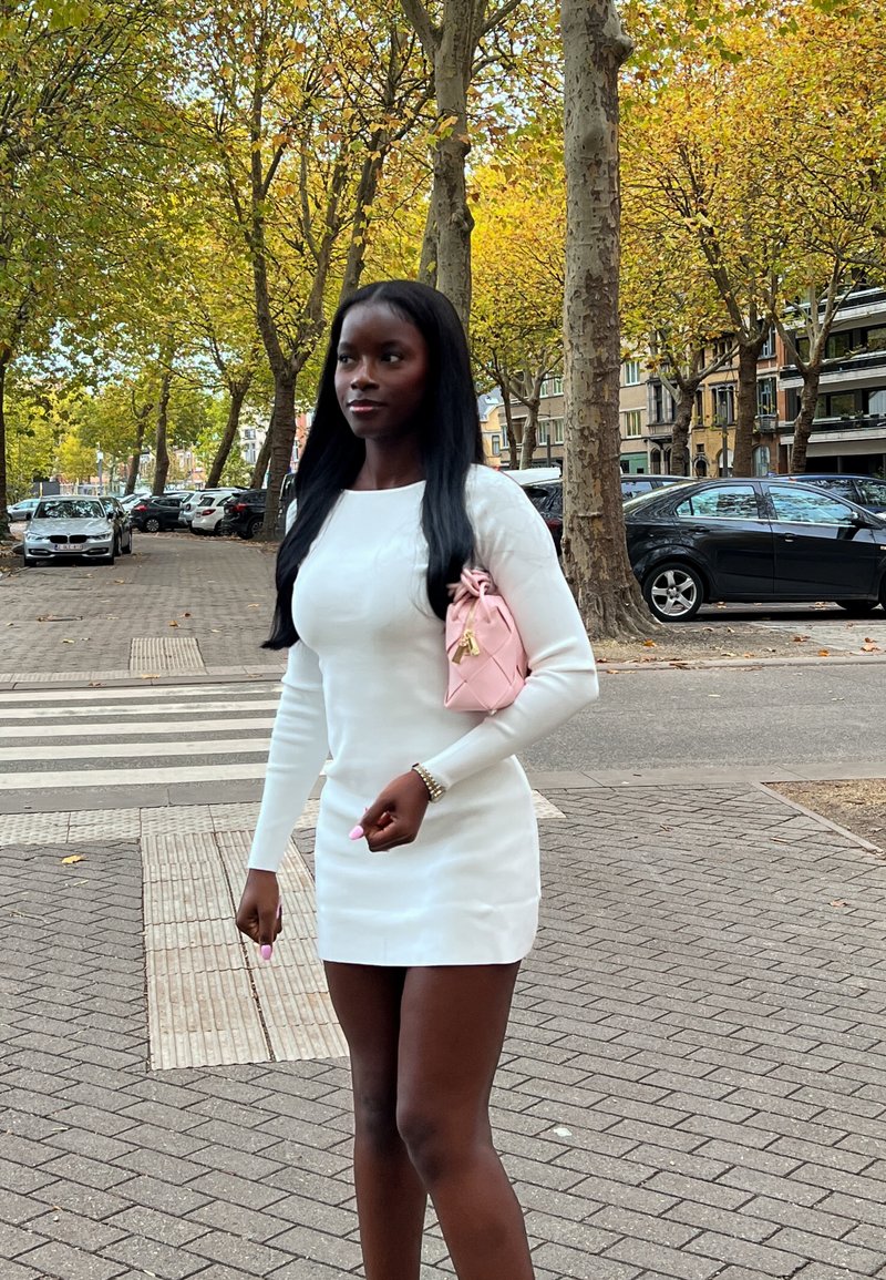 Kvinne i hvit kjole går på fortauet i byen nær en fotgjengerfelt, bærende på en rosa veske, med høsttrær og parkerte biler i bakgrunnen.