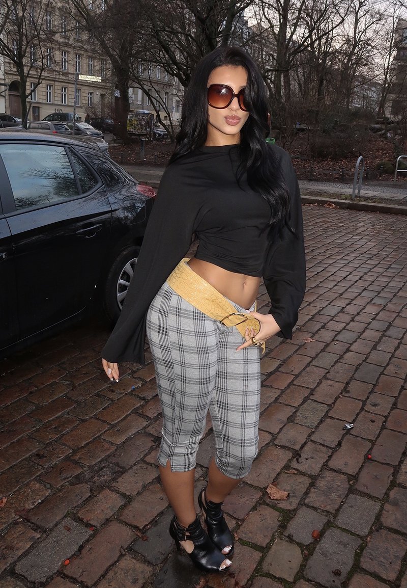 Femme aux longs cheveux noirs et grandes lunettes de soleil, vêtue d'un crop top noir, de pantalons capri à carreaux, d'une ceinture-sac jaune et de bottes noires ouvertes sur un trottoir pavé.