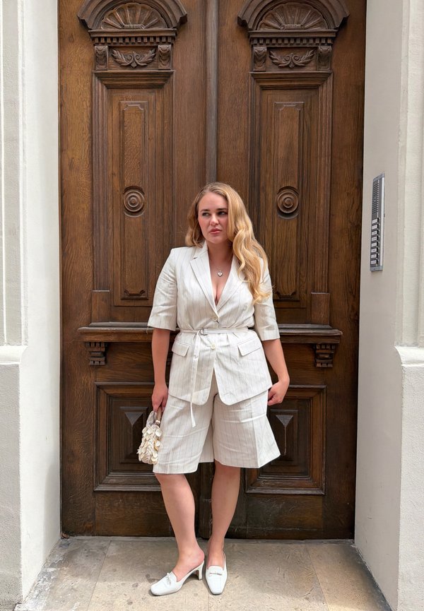 Kvinde med langt blondt hår iført hvidt blazer med striber og shorts står og holder en hvid taske foran en stor udskåret trædobbelt dør.