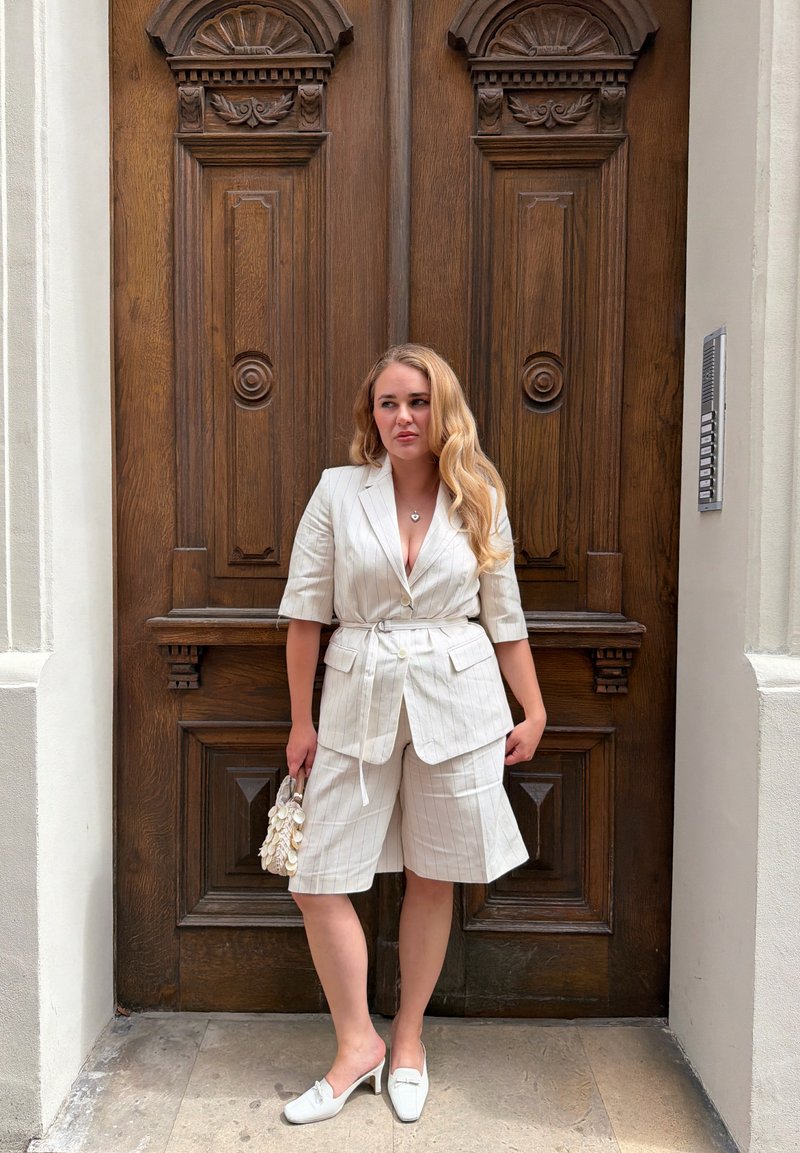 Femeie cu păr blond lung, purtând un sacou alb cu dungi subțiri și pantaloni scurți, stă ținând o geantă albă în fața unei uși mari duble din lemn sculptat.