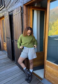 Strikket grønn genser, løs passform; stripete grå shorts; brune støvler opp til knærne; tre-dør bakgrunn; lyst naturlig lys.