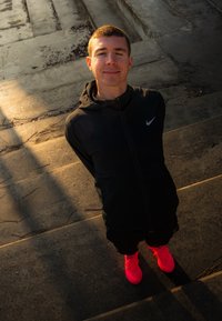 Jeune homme aux cheveux courts, portant une veste Nike noire et des chaussures de course rose vif, debout sur des marches en béton à l'extérieur, sous le soleil.