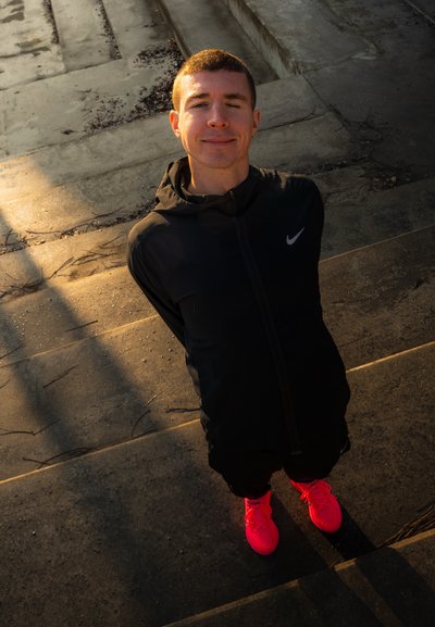 Joven con cabello corto, viste una chaqueta negra de Nike y zapatillas de correr color rosa brillante, de pie en escaleras de concreto al aire libre bajo la luz del sol.