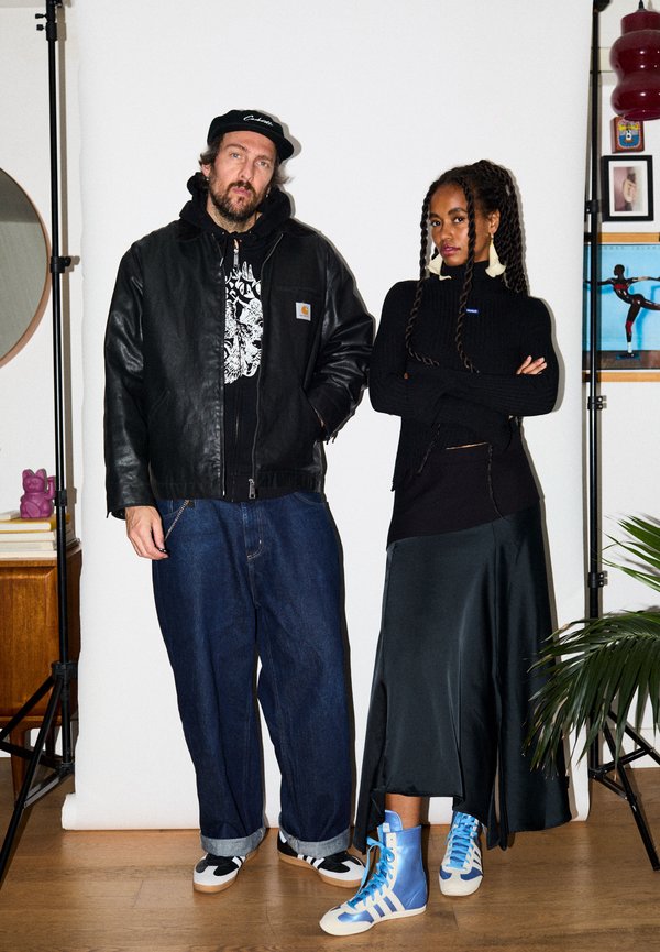 Muž v kožené bundě, čepici a džínách stojí vedle ženy v černém svetru, dlouhé sukni a modrých vysokých teniskách na bílém pozadí.