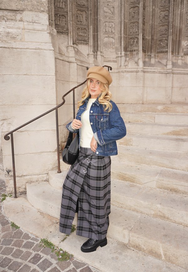 Jeune femme aux cheveux blonds portant un chapeau beige, une veste en jean, un haut blanc, une jupe à carreaux et des chaussures noires, debout sur des marches en pierre près d'un mur orné.