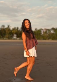 Femme souriante aux longs cheveux bruns marchant pieds nus sur le sable mouillé au coucher du soleil, portant un haut marron et une jupe blanche, avec des arbres en arrière-plan.