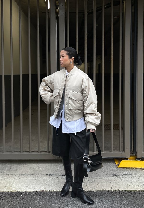 Ženska v bež bombarderski jakni, beli srajci, črnih škornjih do kolen, drži črno torbico, stoji pred sivo kovinsko ograjo.