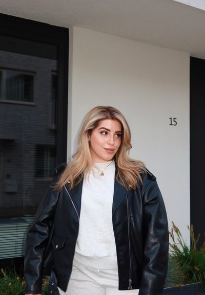 Chaqueta de motorista de cuero negro con acabado brillante, sudadera blanca, pantalones gris claro, de pie cerca de un edificio con el número 15.