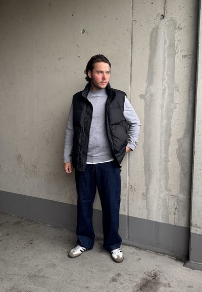 Chaleco negro acolchado sobre un sudadera gris, combinado con jeans oscuros sueltos y zapatillas blancas con acentos grises y negros, de pie contra una pared de concreto.
