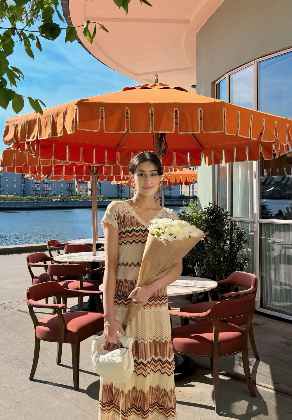Μάξι φόρεμα σε κρεμ απόχρωση με καφέ και σκουροκόκκινες ζιγκ-ζαγκ γραμμές, κρατώντας μια ανθοδέσμη με λευκά λουλούδια, στέκεται κοντά σε πορτοκαλί ομπρέλες και κόκκινες καρέκλες.