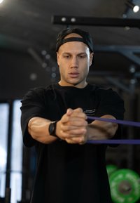 Čierne tričko s krátkym rukávom, fialová odporová guma pevne držaná oboma rukami. Vnútorné fitnescentrum s viditeľnými závažiami v pozadí.