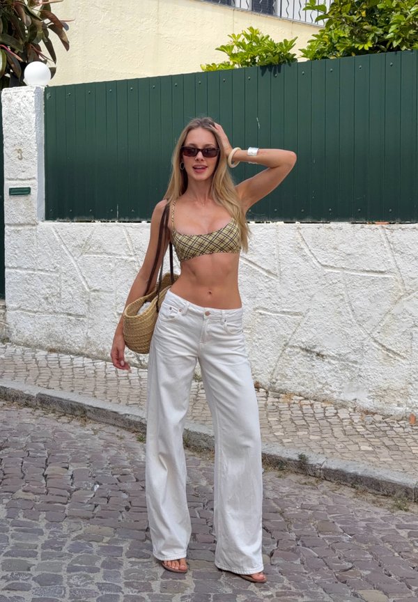 En ung kvinde iført solbriller, ternet cropped top og hvide bukser med vide ben står på en brostensbelagt gade og holder en flettet håndtaske.
