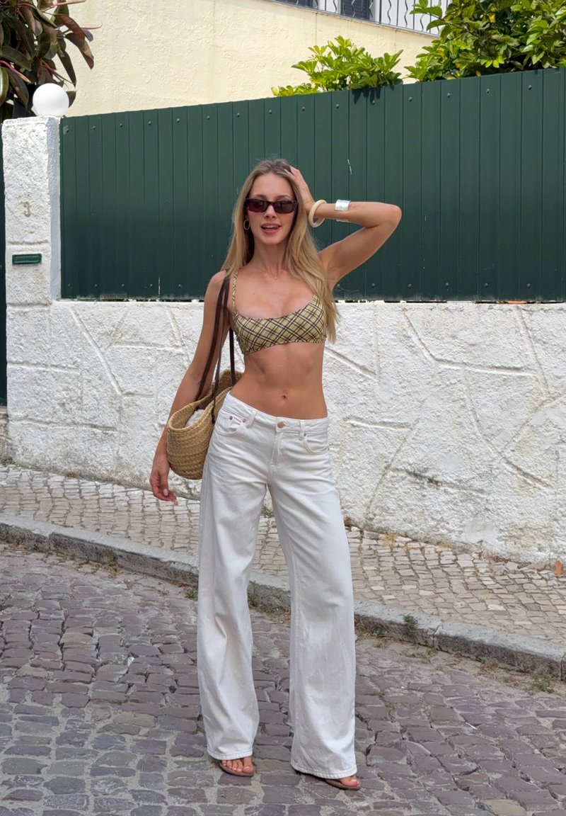 Jeune femme portant des lunettes de soleil, un crop top à carreaux et un pantalon blanc à jambes larges, debout dans une rue pavée, tenant un sac à main tissé.