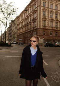 Abrigo negro sobre un suéter azul, camisa azul clara y falda negra. El entorno incluye edificios históricos y una calle con vehículos.