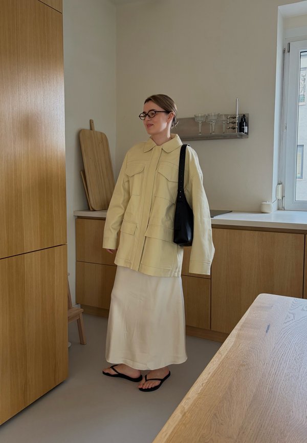 Frau trägt eine beige Jacke und einen Rock mit schwarzen Sandalen und einer Brille, steht in einer modernen Küche mit Holzschränken und einem Fenster.