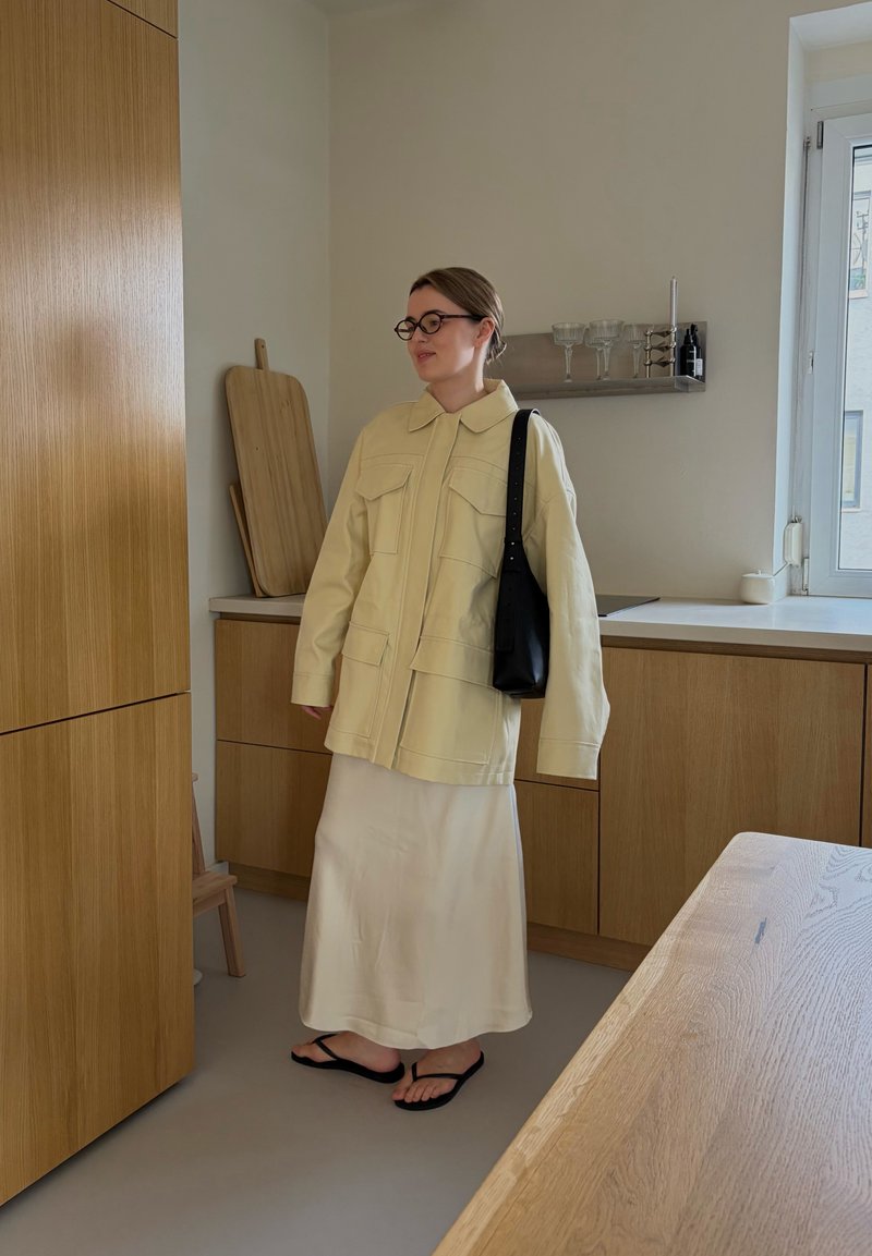 Mujer vestida con chaqueta y falda beige, sandalias negras y gafas, de pie en una cocina moderna con armarios de madera y una ventana.