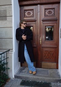 Musta villakangastakki sinisen paidan päällä, vaaleansiniset farkut ja ruskeat kengät. Seisoo puisen, koristeellisin yksityiskohdin varustetun kaksiosaisen oven vieressä.